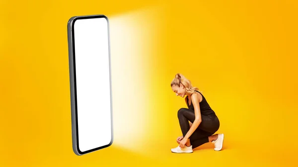 Sportliche Frau schnürt Schuhe in der Nähe des großen Smartphone-Bildschirms, gelber Hintergrund — Stockfoto