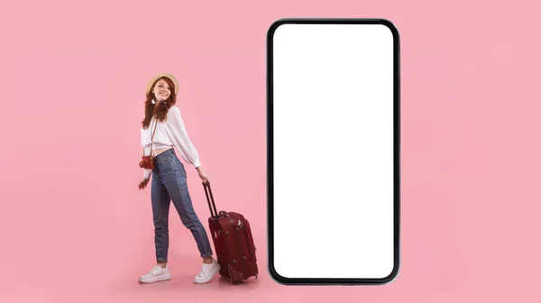 Turista femenina cerca de la pantalla del teléfono posando con la maleta, fondo rosa — Foto de Stock