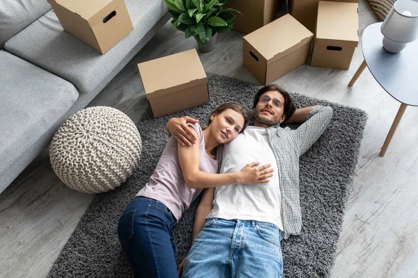 Уставшие улыбающиеся европейские тысячелетия муж и жена радуются покупке новой собственной квартиры, лежат на полу — стоковое фото