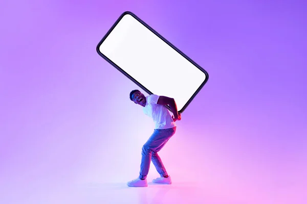 Ελκυστικό milennial μαύρο άντρα που μεταφέρουν μεγάλο βαρύ smartphone με λευκή οθόνη στην πλάτη του σε νέον φως — Φωτογραφία Αρχείου