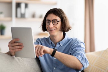 Boş zaman kavramı. Mutlu genç adam dijital tablet kullanıyor, internetten alışveriş yapıyor, kanepede dinlenirken internette geziniyor.