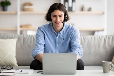 Kulaklık takan mutlu genç adam, online toplantı yapıyor ya da Webinar 'a katılıyor, evden çalışıyor.