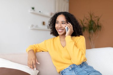 Neşeli genç Afrikalı Amerikalı kadın akıllı telefonla konuşuyor, evdeki kanepede oturuyor.