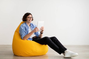 Armut koltukta dijital tabletle rahatlayan genç adam, modern aletler kullanan erkek öğrenci, boş alan.