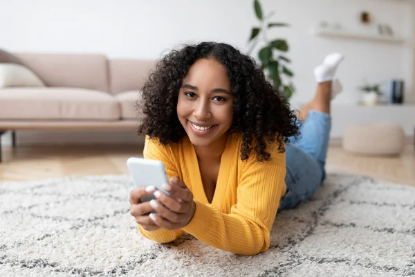 Felice giovane donna nera utilizzando il telefono cellulare, avendo chiamata online, sdraiato sul pavimento, pubblicazione nei social media a casa — Foto Stock