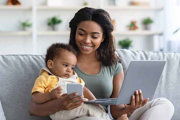 Vrouw met laptop en mobiele telefoon thuis met kind in de armen — Stockfoto