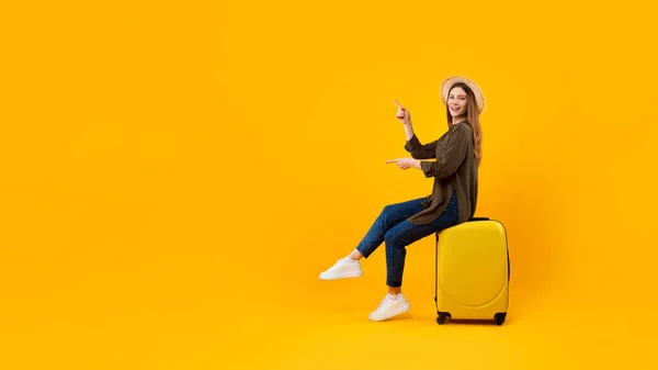 Weibliche Touristin zeigt mit Fingern auf Koffer sitzend, gelber Hintergrund — Stockfoto