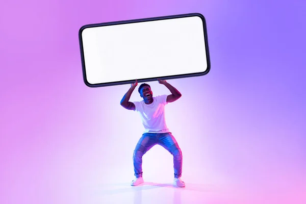 Hezký mladý černoch drží obrovský těžký smartphone s prázdnou bílou obrazovkou v neonovém světle, mockup — Stock fotografie