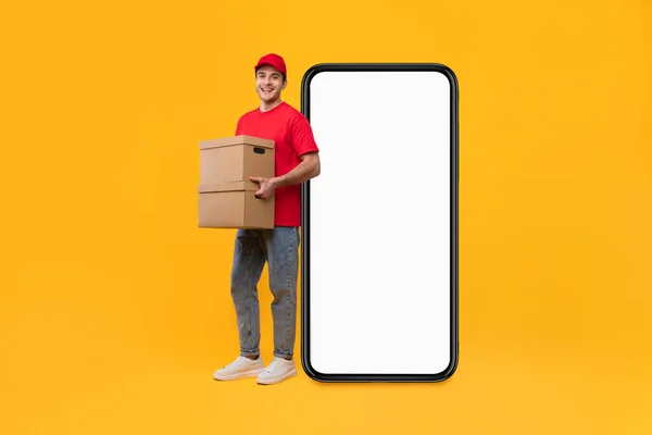 Entregador perto de tela grande smartphone segurando caixas de papelão, fundo amarelo — Fotografia de Stock