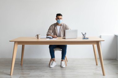 Koruyucu maskeli bir adam merkez büroda bilgisayar üzerinde çalışıyor.