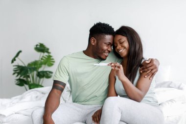 Mutlu Afrikalı genç Amerikalı kadın ve erkeğin yatak odasındaki beyaz yatakta hamilelik testine bakmasına sevindim.
