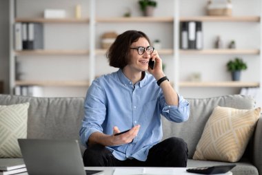 Her zaman görüşüyoruz. Genç serbest çalışan adam cep telefonuyla konuşuyor ve bilgisayarla evden uzaktan çalışıyor, kanepede oturuyor.