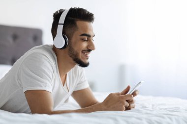 Boş Zaman Teknolojileri. Kulaklıklı Orta Doğulu Adam Yatakta Akıllı Telefon Kullanıyor