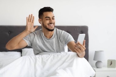 Yatakta akıllı telefonlu neşeli bir Arap arkadaşlarıyla video görüşmesi yapıyor.