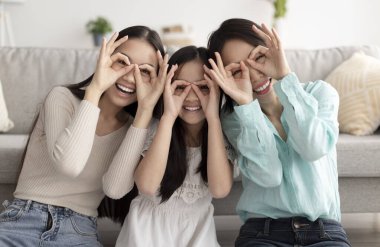 Annesi ve büyükannesiyle birlikte küçük Asyalı kız eğleniyor, aptalca davranıyor, evde gözlük jesti yapıyor.