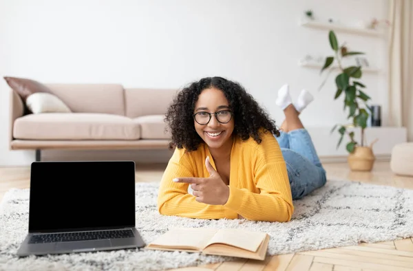 Comprimento total de feliz jovem mulher negra deitada no chão, apontando para a tela do laptop pc com mockup para o design do site — Fotografia de Stock