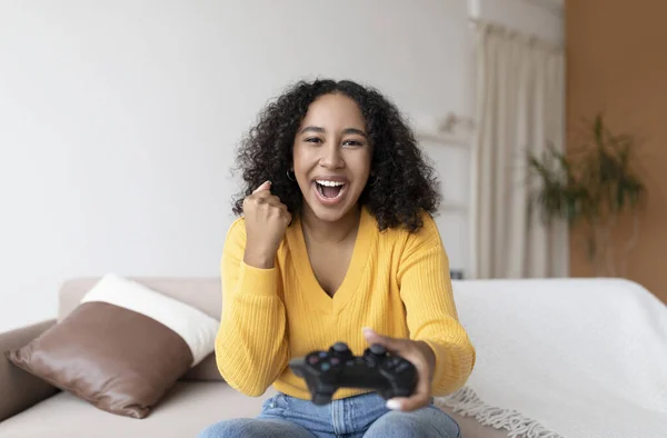 Eccitato giovane donna afroamericana con joystick fare SI gesto, vincente videogioco, seduto sul divano a casa — Foto Stock