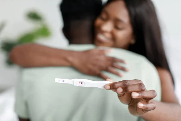 Радостная улыбающаяся чернокожая женщина обнимающая мужчину показывает тест на беременность в интерьере дома, крупным планом, размытым — стоковое фото