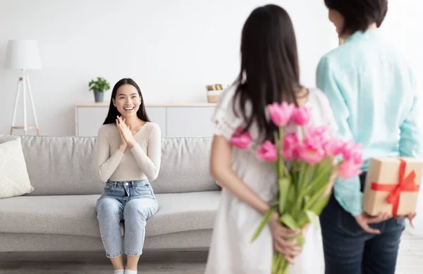 Liten asiatisk flicka med sin mormor gömmer gåva och blommor för ung kvinna bakom ryggen på dem hemma. Familjesemester och firande koncept — Stockfoto