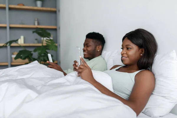 Smějící se mladý černošky chlap a žena chatování na smartphonech ležet na bílé posteli v ložnici — Stock fotografie