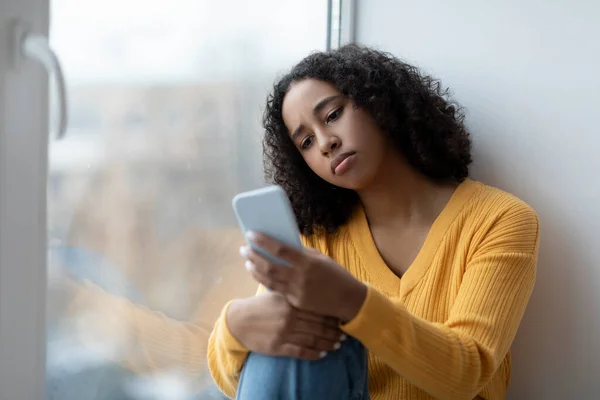 Depressieve jonge Afro-Amerikaanse vrouw leest schokkend nieuws op smartphone, zit in de buurt van venster thuis, kopiëren ruimte — Stockfoto