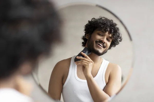 Omhändertagande koncept. Stilig ung indian man tittar på spegel och rakning skägg med trimmer eller elektrisk rakapparat — Stockfoto