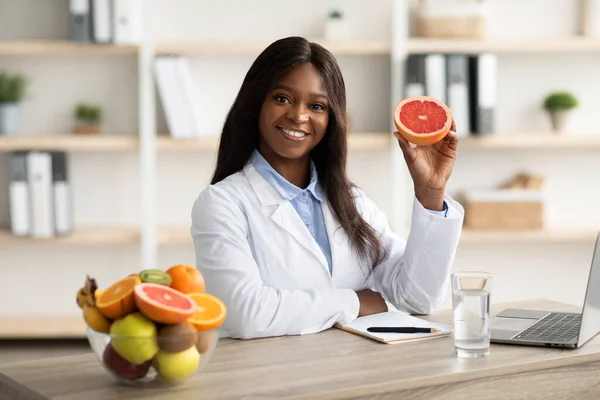 Amistosa dietista femenina negra sosteniendo pomelo y sonriendo a la cámara, ofreciendo consultas de pérdida de peso en línea —  Fotos de Stock