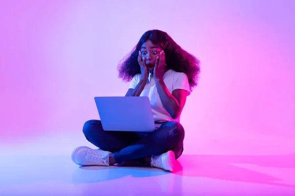 Lunghezza completa della donna afroamericana scioccata che utilizza il computer portatile, aprendo la bocca in sorpresa nella luce al neon — Foto Stock