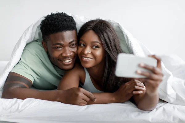 Χαρούμενα νεαρά μαύρα αρσενικά και θηλυκά αγκαλιάζει κάτω από τα καλύμματα λάβει selfie και να χαλαρώσετε στο κρεβάτι στο υπνοδωμάτιο — Φωτογραφία Αρχείου
