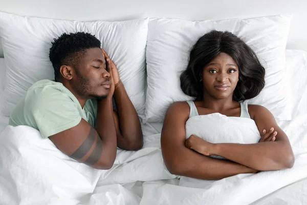 Δυσαρεστημένη θυμωμένη αστεία νεαρή Αφροαμερικανή που υποφέρει από ροχαλητό, κοιμισμένο άντρα. — Φωτογραφία Αρχείου