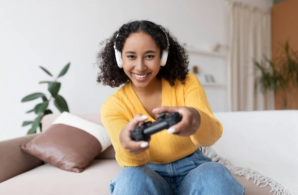 Mujer negra milenaria feliz en auriculares usando joystick para jugar videojuego en el sofá en casa. Pasatiempos domésticos —  Fotos de Stock