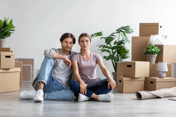 Graves millennial europeo chico y señora sentado en el suelo entre cajas y plantas en la habitación, tomar un descanso —  Fotos de Stock