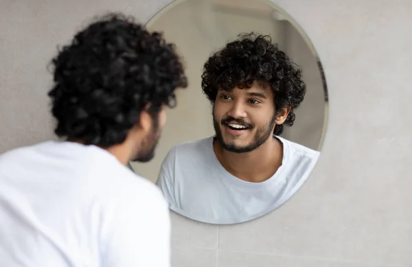 Retrato de un hombre indio feliz mirando su reflejo en un espejo redondo en la pared, un joven parado en el baño —  Fotos de Stock