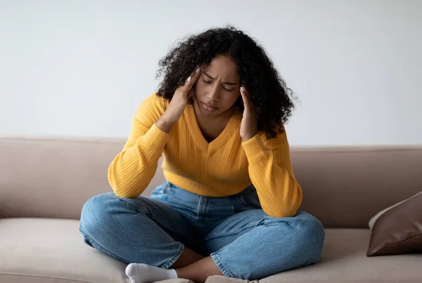 Joven mujer negra con depresión tocando sus sienes, sintiéndose estresada en el sofá en casa — Foto de Stock