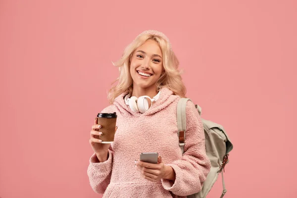 Hezká dáma pózuje s batohem, drží kávu jít a mobilní telefon, usmívá se na kameru přes růžové pozadí studia — Stock fotografie
