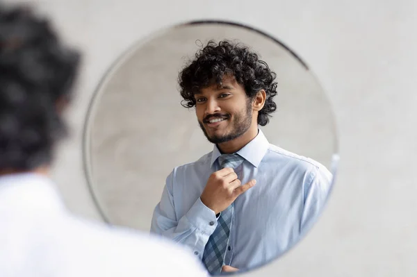 Hombre de negocios indio guapo poniéndose la corbata y mirando su reflejo en el espejo, de pie en el baño —  Fotos de Stock