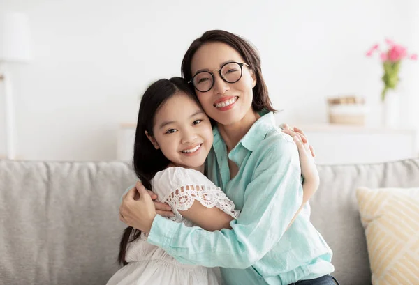 Feliz niña asiática y su abuela cariñosa abrazando y mirando a la cámara en el sofá en casa — Foto de Stock