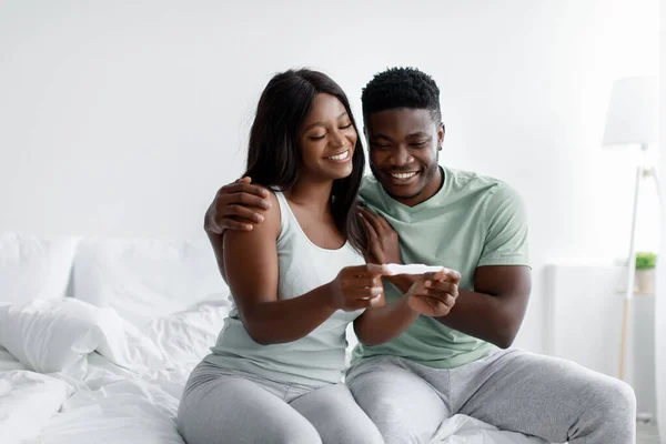 Χαρούμενη νεαρή Αφροαμερικανή γυναίκα δείχνει θετικό τεστ εγκυμοσύνης στον άντρα της στο εσωτερικό της κρεβατοκάμαρας. — Φωτογραφία Αρχείου