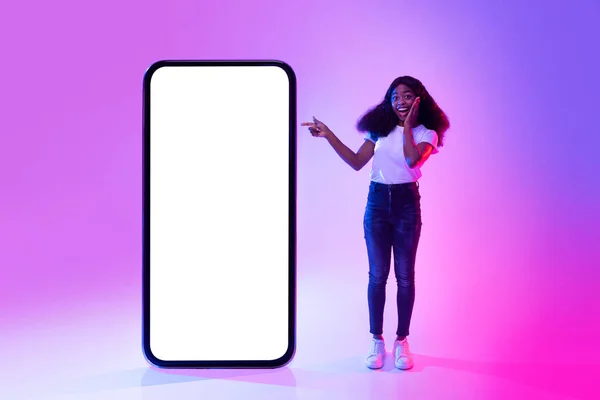 Femme noire excitée pointant du doigt un gros téléphone portable avec écran blanc vide, faisant la promotion d'une nouvelle application ou d'un nouveau site Web au néon, maquette — Photo