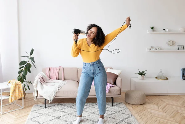 Giovane donna afroamericana positiva che usa l'asciugacapelli come microfono, canta e balla, si diverte a casa — Foto Stock