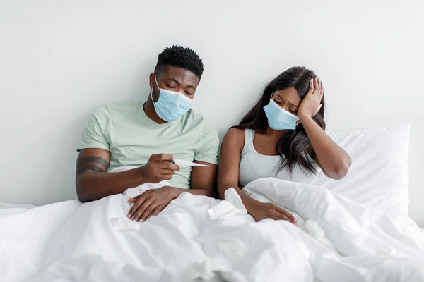 Deprimerad ung svart dam och kille i skyddsmasker titta på termometer på vit säng i sovrummet — Stockfoto