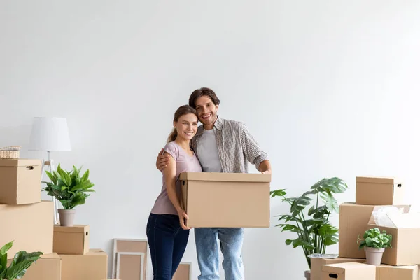 Glimlachende gelukkige duizendjarige blanke man en vrouw met kartonnen doos knuffelen in hun eigen appartement — Stockfoto