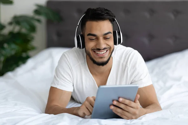 Feliz chico árabe con auriculares inalámbricos relajante con tableta digital en la cama — Foto de Stock