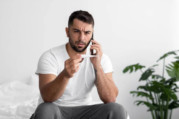 Infeliz europeo millennial chico sufre de problemas de salud, mira el termómetro, llamada por teléfono sentarse en la cama — Foto de Stock