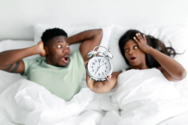 Sokkolt Y-generációs fekete férfi és nő nyitott szájjal mutatják ébresztőóra, fekszik a fehér ágy a hálószobában — Stock Fotó