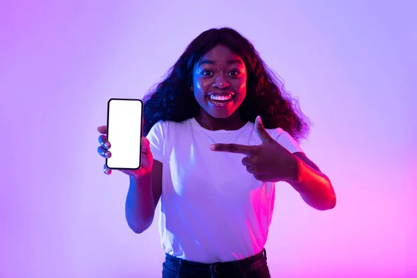 Retrato de alegre joven hembra negra apuntando al teléfono inteligente con pantalla blanca vacía en luz de neón — Foto de Stock