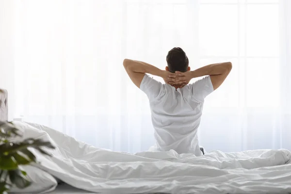 Guten Morgen. Mann mit Händen hinter dem Kopf auf Bett sitzend, Rückansicht — Stockfoto