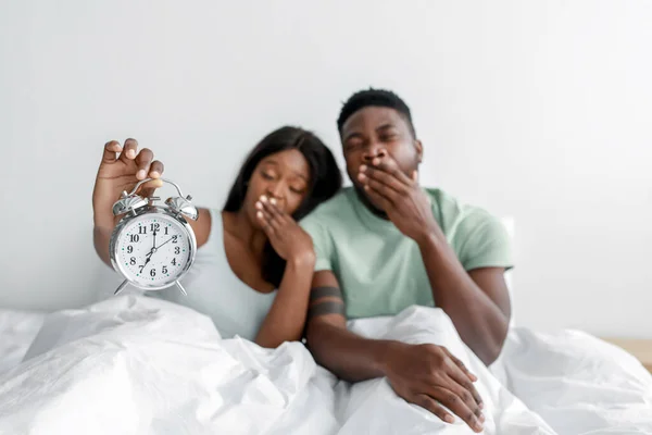 Сплячий молодий афроамериканець і жінка прокидаються вночі, позіхають, сідають на ліжко в спальні, дружина тримає будильник. — стокове фото