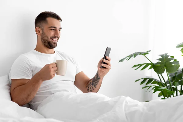Χαρούμενος καυκάσιος ελκυστικός άντρας σε λευκό t-shirt με φλιτζάνι καφέ στο κρεβάτι στην κρεβατοκάμαρα — Φωτογραφία Αρχείου