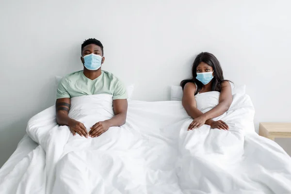 Depresión triste joven negro chico y mujer en máscaras protectoras sentarse a distancia en la cama en el interior del dormitorio —  Fotos de Stock